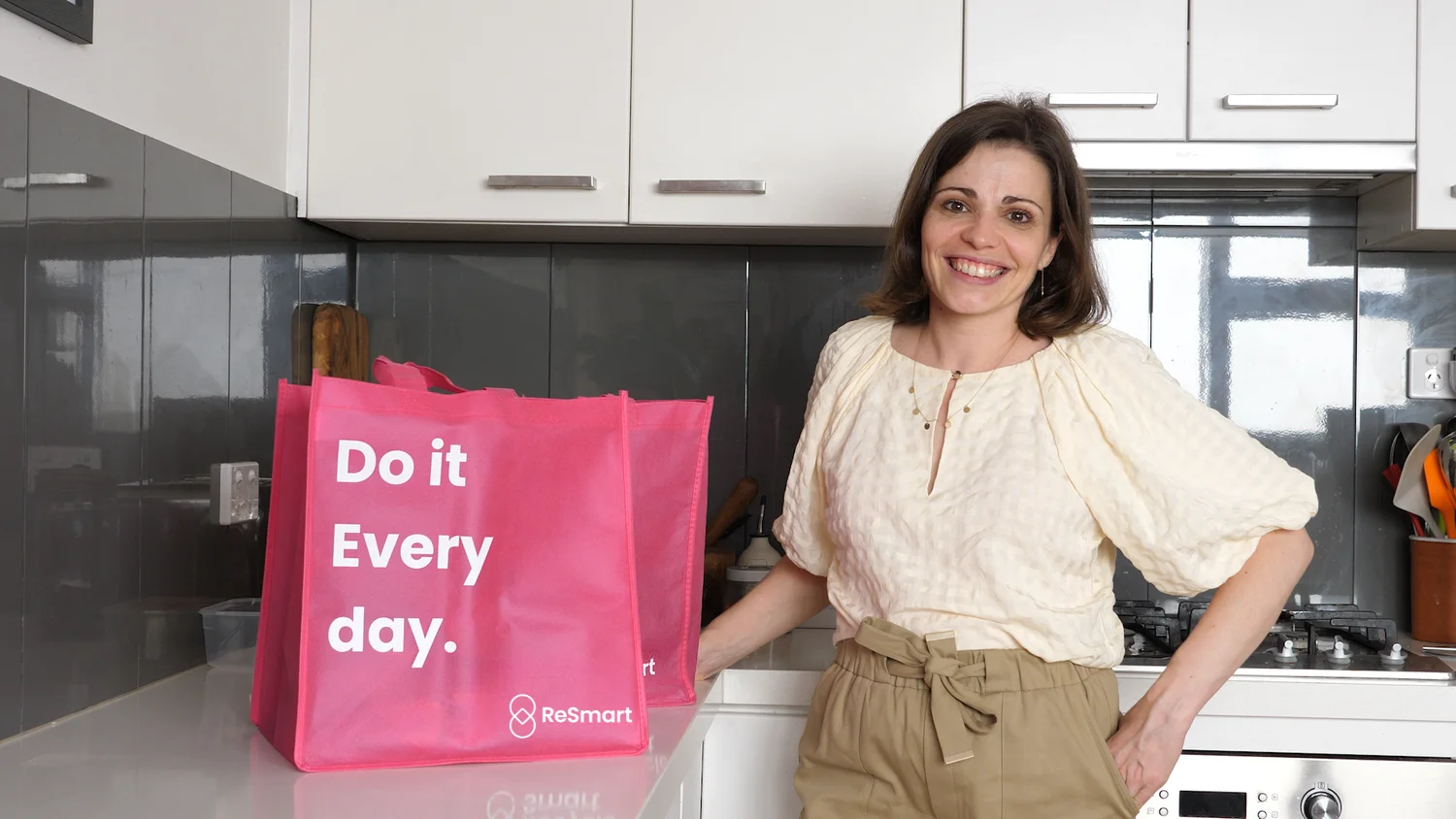 A woman smiling in a kitchen standing beside a pink reusable shopping bag with white text saying 'Do it Every day.'