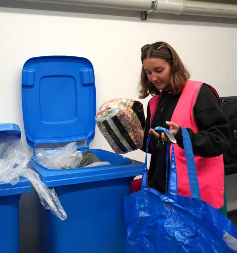 Woman in a pink safety vest placing a wrapped package into a blue recycling bin