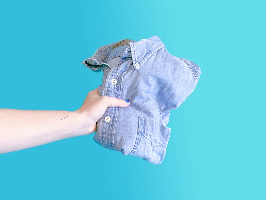Hand holding a folded denim shirt against a turquoise background