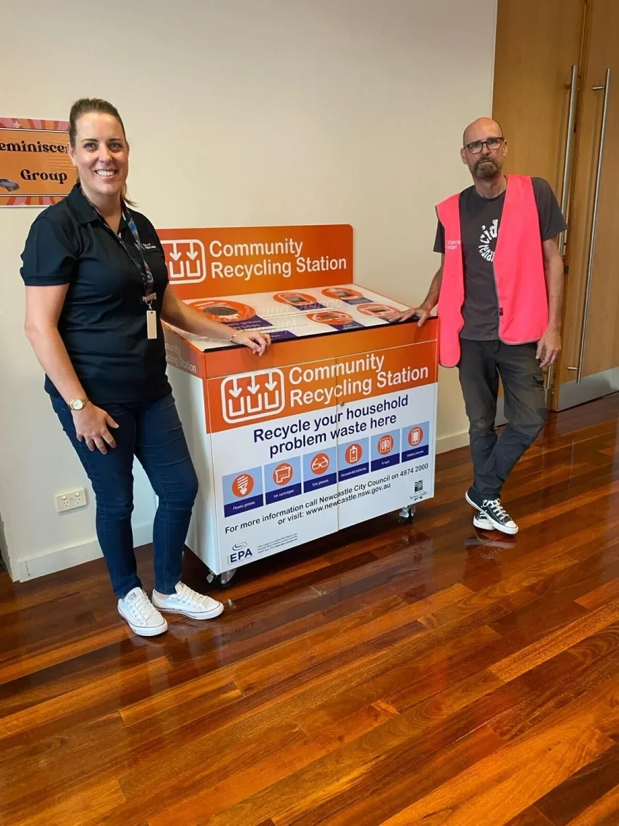 Two people standing beside a community recycling station with orange signage